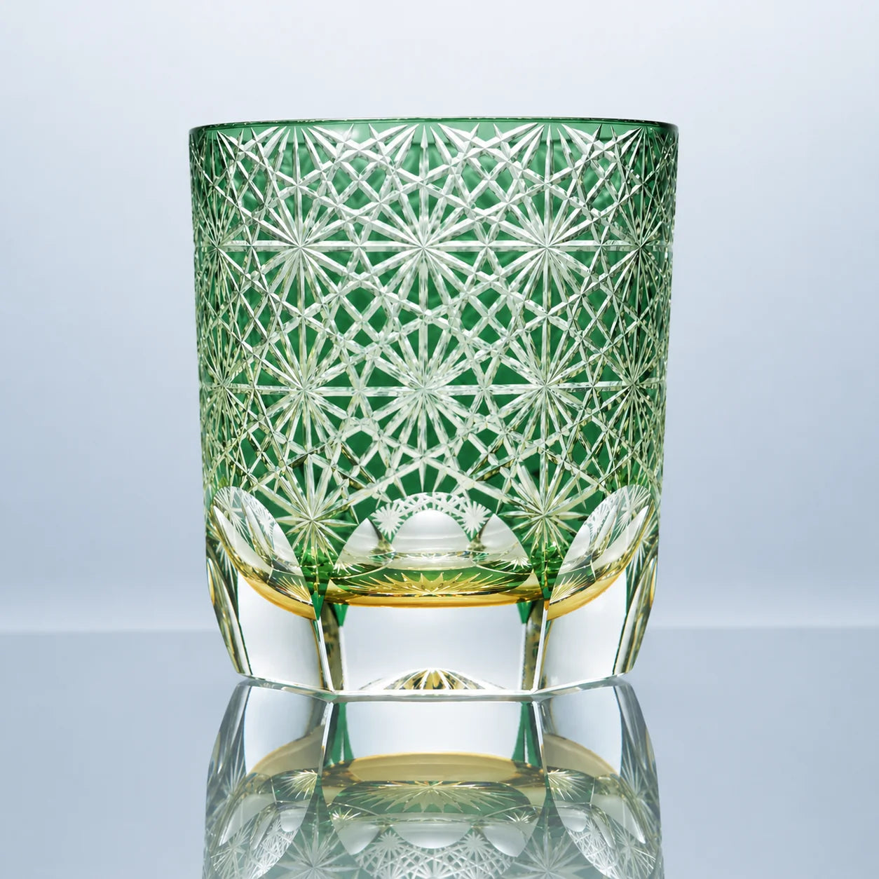 A small green hand-cut Edo Kiriko crystal whiskey glass featuring daisy-inspired starburst patterns, a soft emerald tone, and a polished clear faceted base in a reflective studio setting.