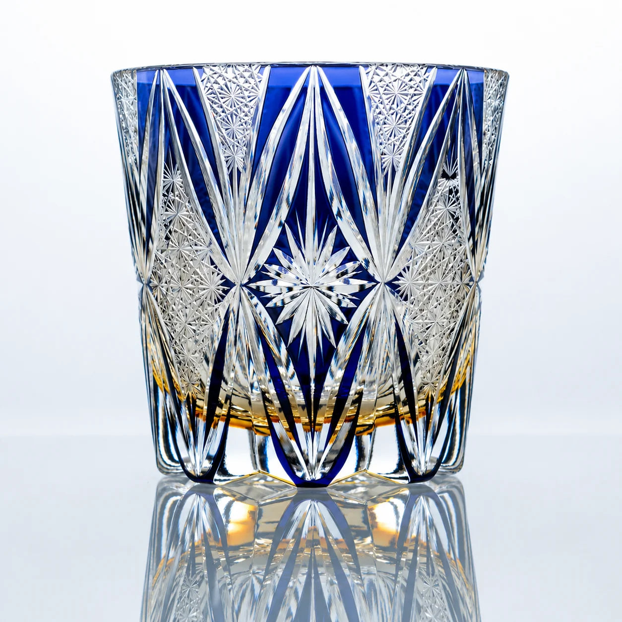 A hand-cut Edo Kiriko crystal whiskey glass featuring a vivid blue upper body, clear geometric cut details, a central starburst motif, and a polished reflective studio presentation.