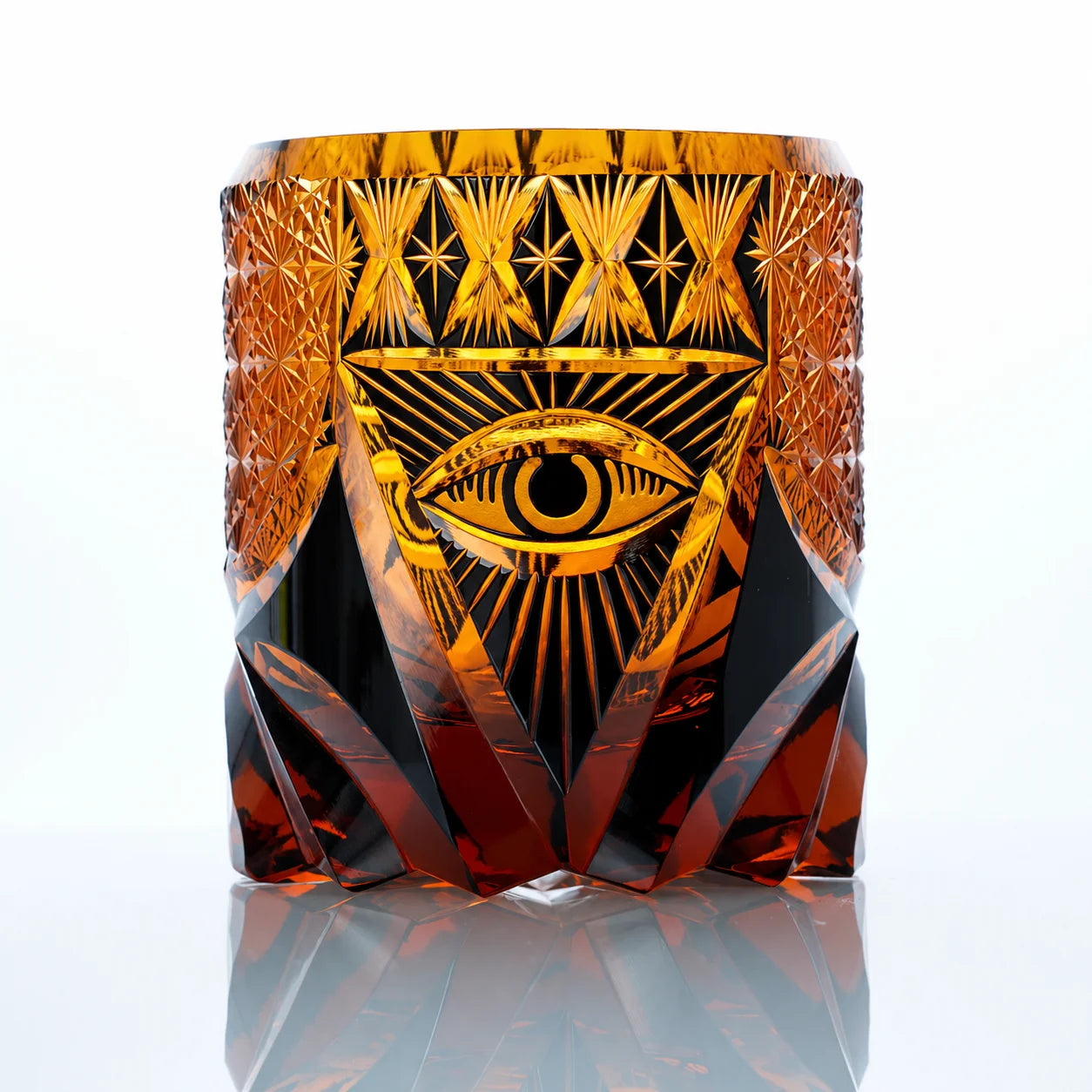 An amber hand-cut Edo Kiriko whiskey glass featuring a symbolic eye motif, radiant geometric crystal cuts, and a reflective studio presentation.