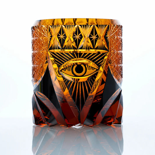 An amber hand-cut Edo Kiriko whiskey glass featuring a symbolic eye motif, radiant geometric crystal cuts, and a reflective studio presentation.