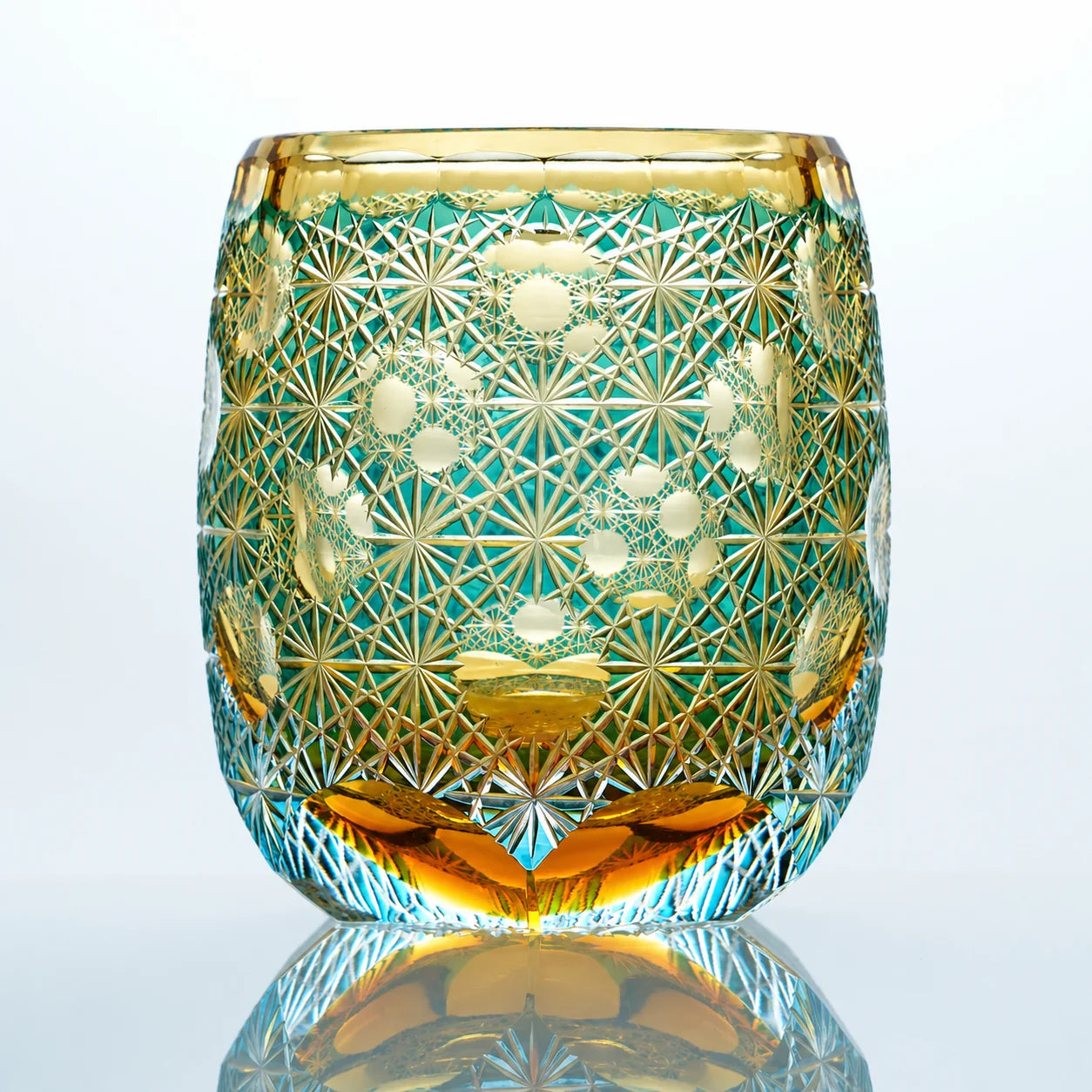 An aqua amber hand-cut Edo Kiriko whiskey glass featuring teal-green crystal tones, warm amber accents, intricate starburst patterns, and a reflective studio presentation.