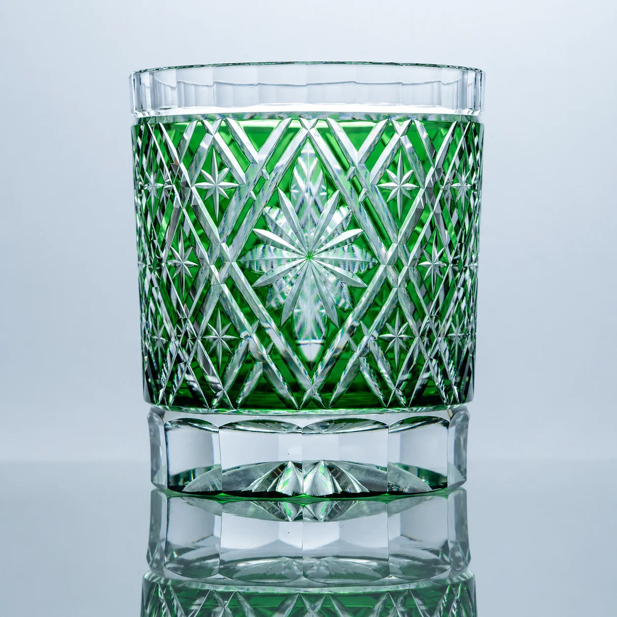 An emerald hand-cut Edo Kiriko whiskey glass featuring a radiant starburst center, geometric lattice crystal cuts, a clear faceted base, and a polished reflective studio presentation.