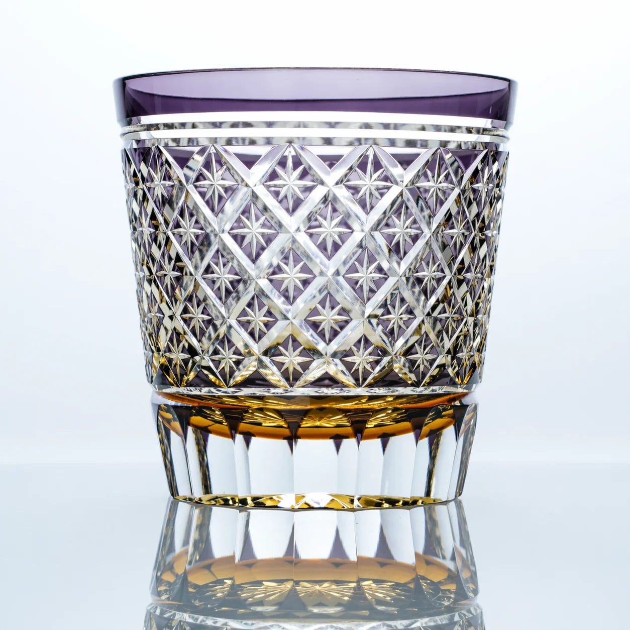 A hand-cut Edo Kiriko whiskey glass featuring a lavender-purple upper band, intricate star lattice cutwork, a faceted crystal body, and a warm golden base in a clean reflective studio setting.