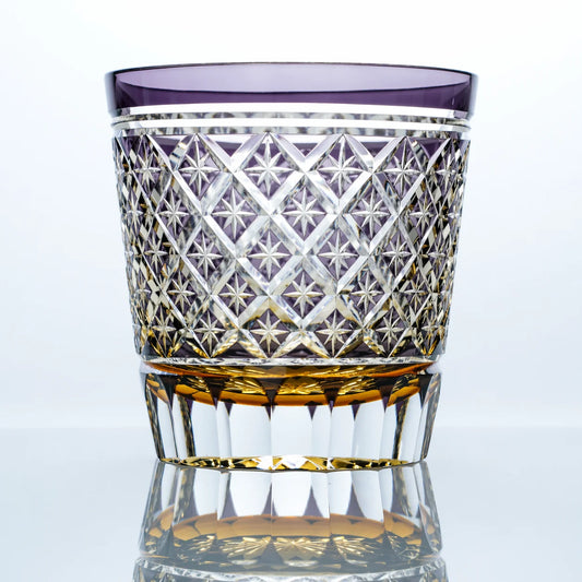A hand-cut Edo Kiriko whiskey glass featuring a lavender-purple upper band, intricate star lattice cutwork, a faceted crystal body, and a warm golden base in a clean reflective studio setting.