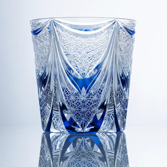 A front view of a sapphire blue hand-cut Edo Kiriko whiskey glass featuring sweeping veil-like crystal facets, dense starburst textures, and a reflective studio presentation.