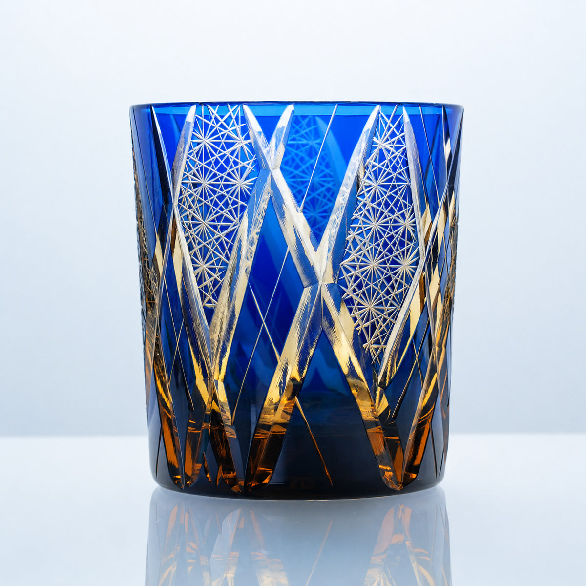 A hand-cut Edo Kiriko whiskey glass in vivid sapphire-blue crystal, featuring warm amber-gold accents, tall interlacing cuts, and intricate geometric crystal detailing.